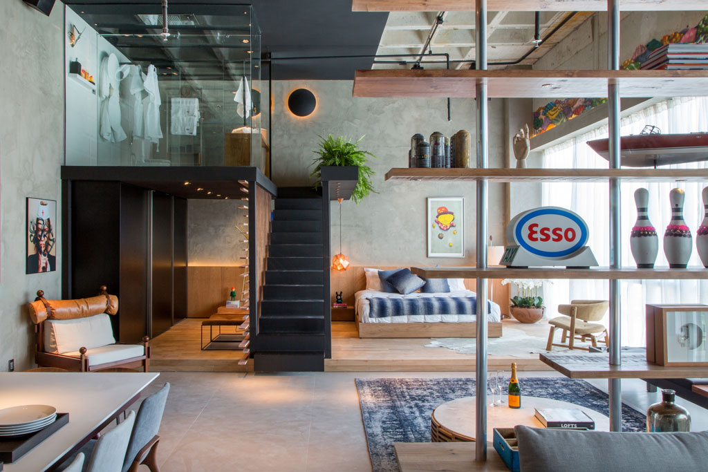 CASA COR RIO 2014. Ambiente: Loft do Solteiro. Alexandre Gedeon e Hugo Schwartz removeram o rebaixo de gesso existente para deixar aparente a estrutura da laje de concreto, além de tirar proveito do pé direito de quase 6 m. Assim, o mezanino em aço passa a abrigar o banheiro. No nível inferior, ambientes público e privado são diferenciados pelo piso: frio para a área social (cozinha, estar e home theater) e de madeira no quarto e no closet. O conforto e o design estão aliados na poltrona Tonico, de Sérgio Rodrigues, e na cadeira Joy, de Jader de Almeida. As mesas Fafá G e Fafá M são de Rejane Carvalho. Em primeiro plano, a estante que divide estar e home theater, criação da dupla, é toda feita com tábuas de pinho e estrutura tipo andaime em ferro.