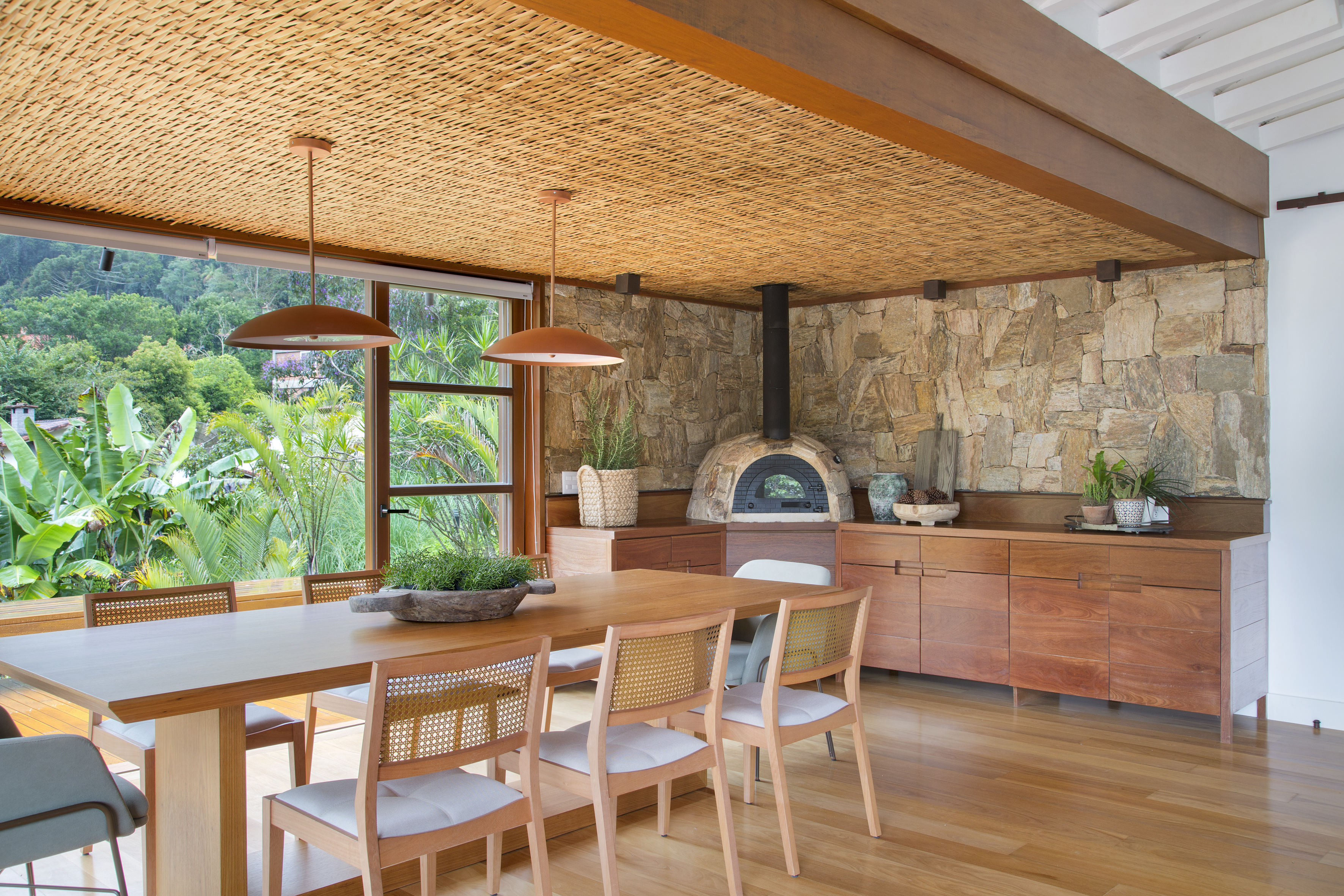 Suíte de casa de campo fica em bangalô suspenso no meio da natureza. Projeto de Escala Arquitetura. Na foto, sala de jantar com teto de fibra e vista para o jardim. Churrasqueira e parede de pedra.