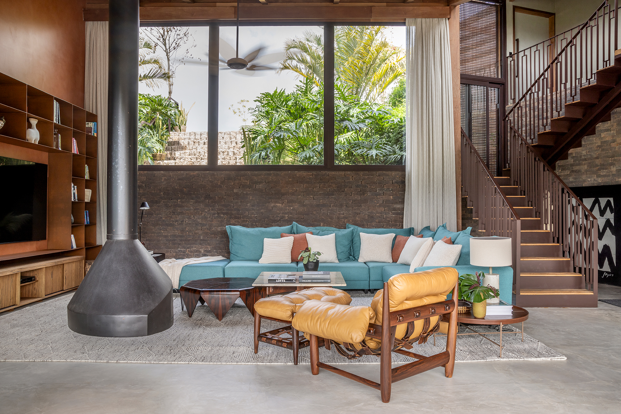 Casa sustentável e integrada à natureza fica em frente ao campo de golfe. Projeto de In House Arquitetura. Na foto, sala de estar com sofá azul e lareira. Poltrona Mole e escada.