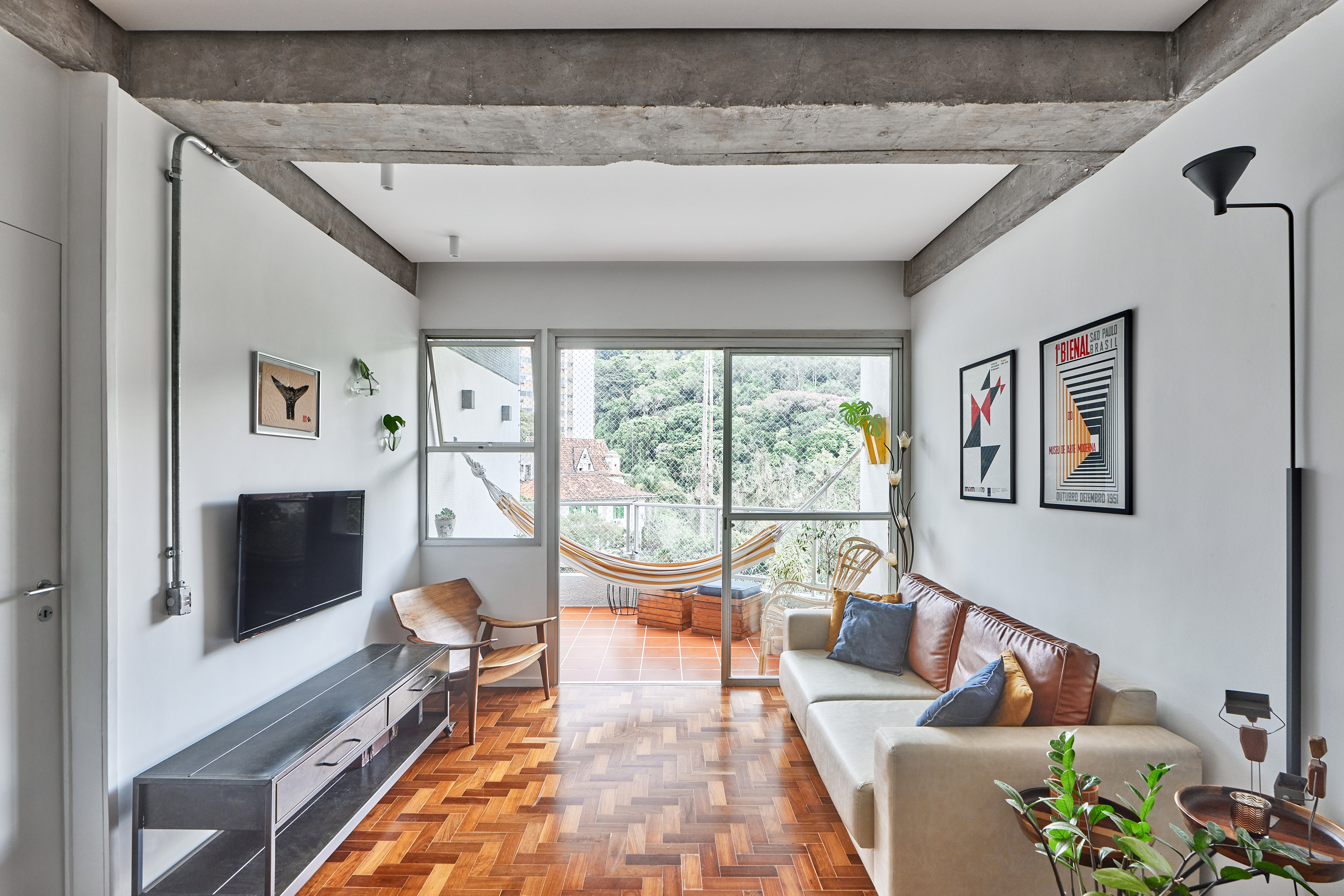 Apartamento de 98 m² ganha décor contemporâneo, mas com toques de brutalismo. Projeto de Hugo Rapizo. Na foto, sala com sofá, quadro, piso de taco e varanda com rede.