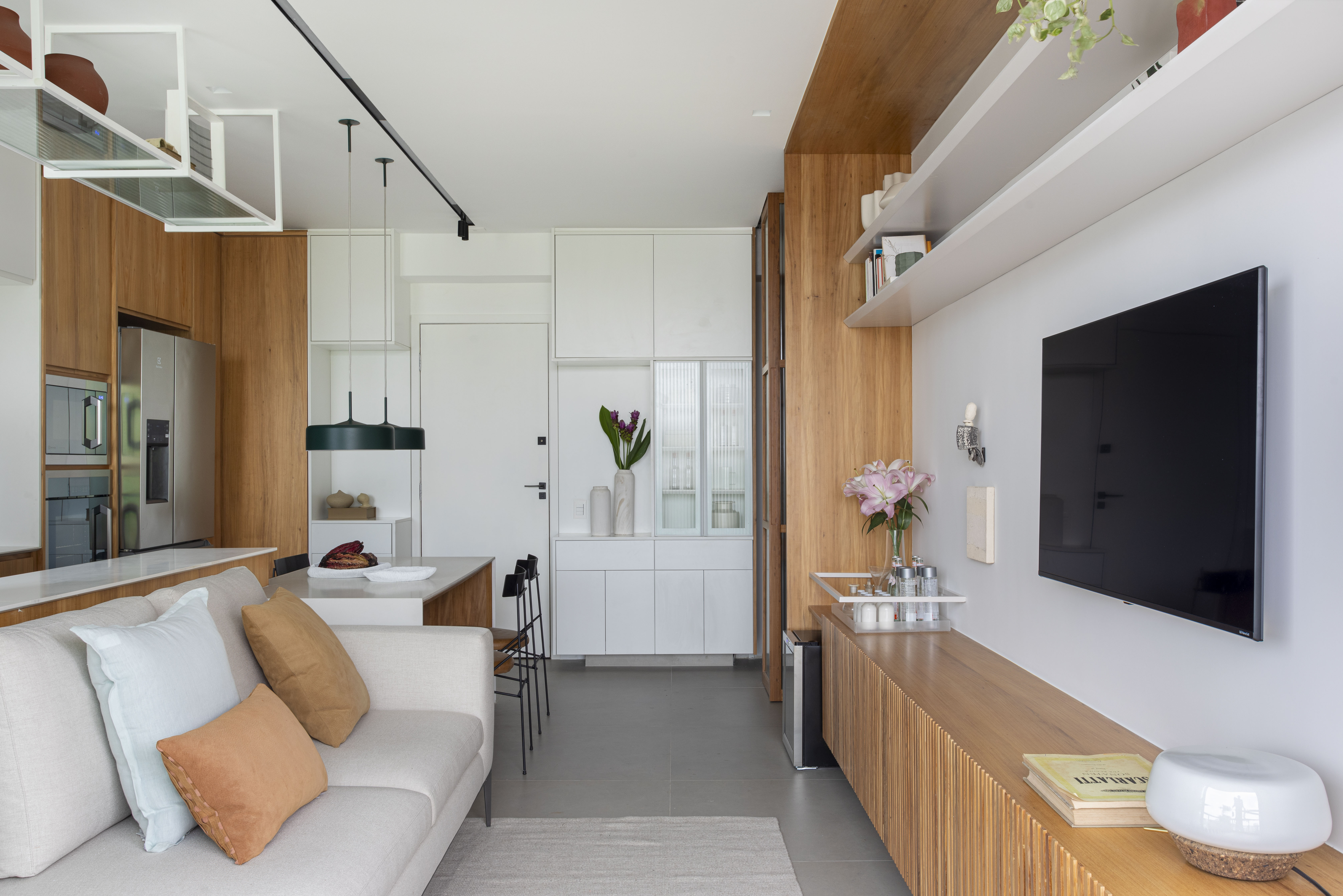 Branco e madeira criam décor cozy em apartamento de 70 m². Projeto de Rafael Ramos. Na foto, sala de tv com aparador ripado e prateleiras. Cozinha integrada.