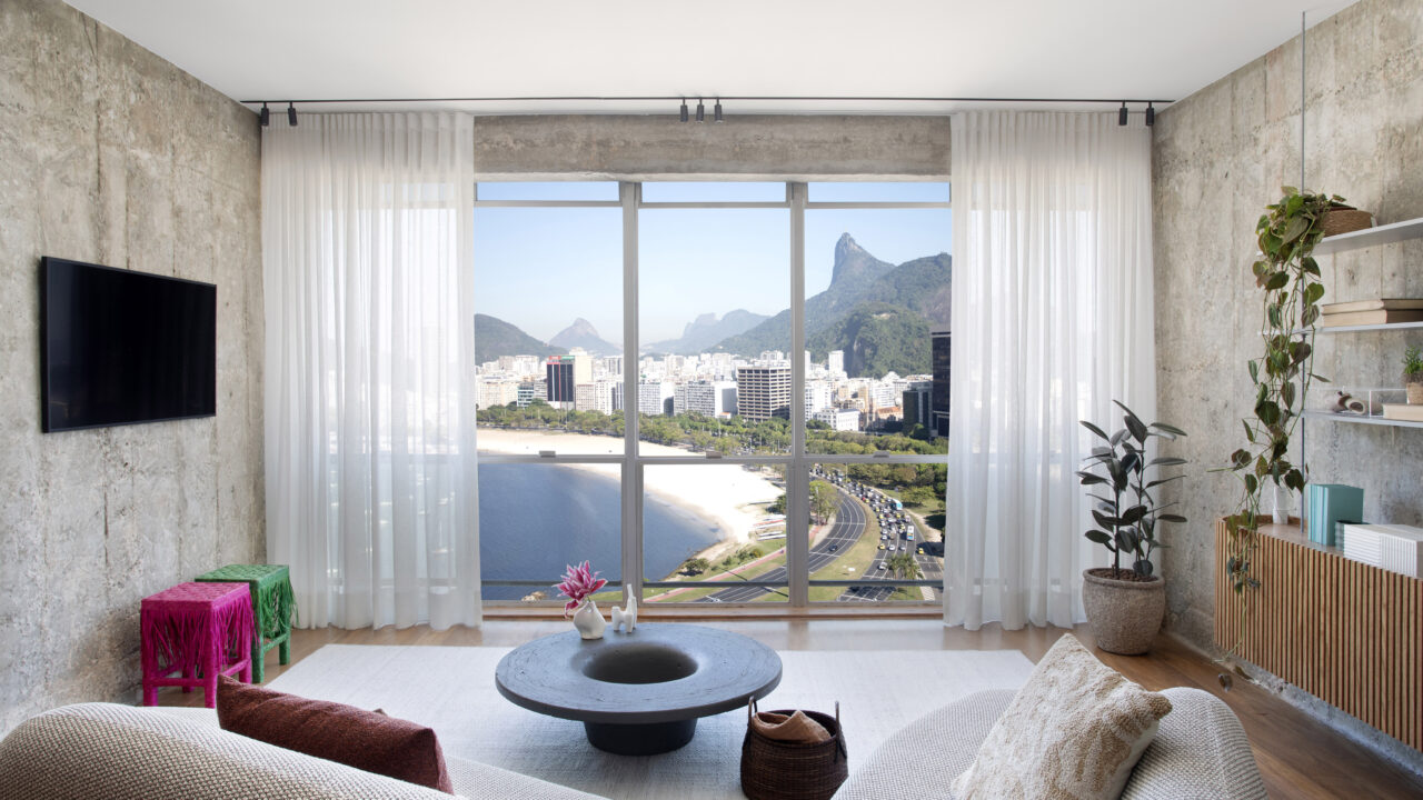 Rafael Ramos emoldura a paisagem carioca neste apartamento de 98 m². Na foto, sala com parede de concreto, vista para a praia e sofá.