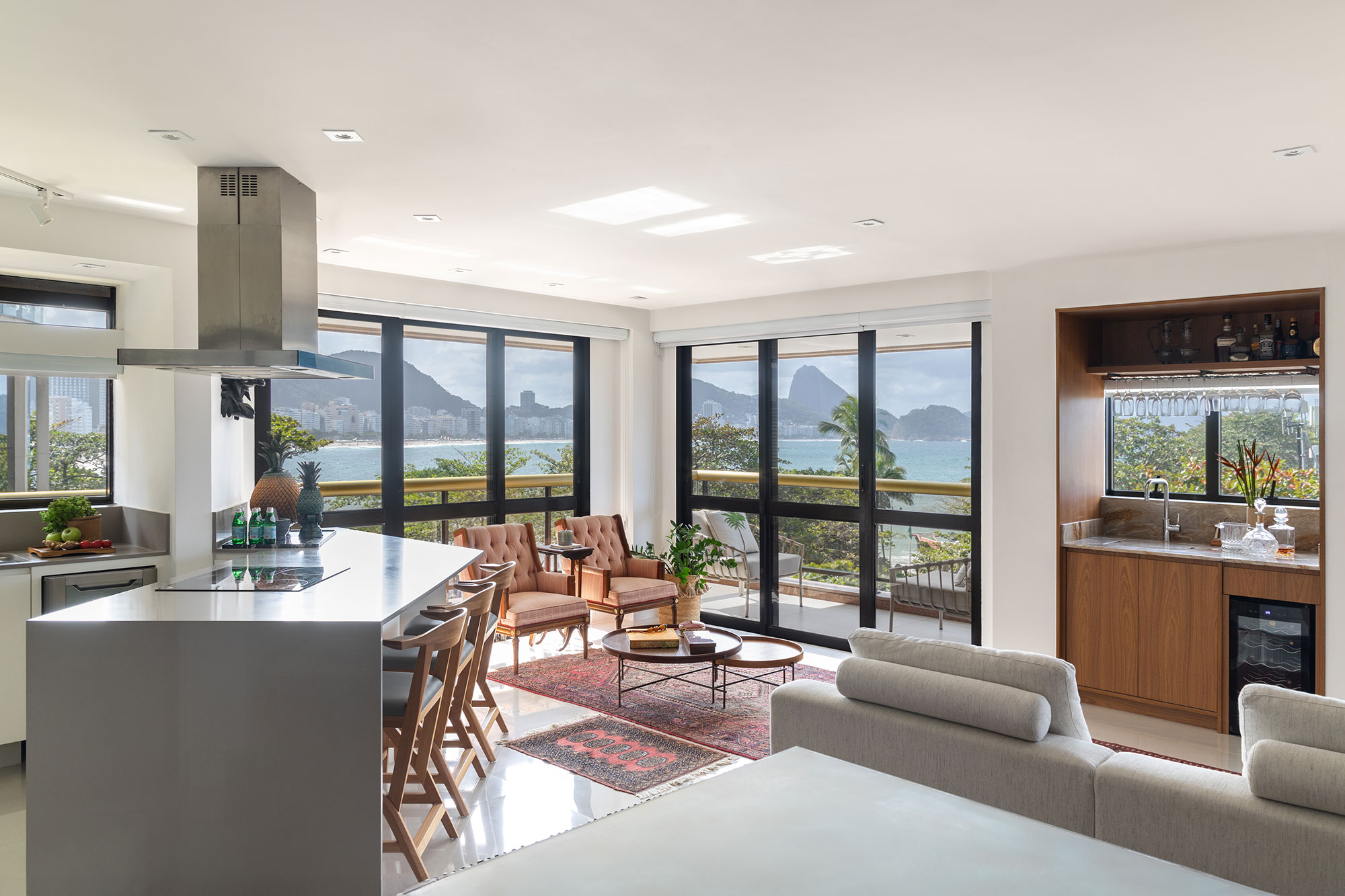 Vivian Reimers une dois apês para criar projeto com vista para o Pão de Açúcar. Na foto, cozinha com ilha e banquetas. Sala com tapete e poltronas.