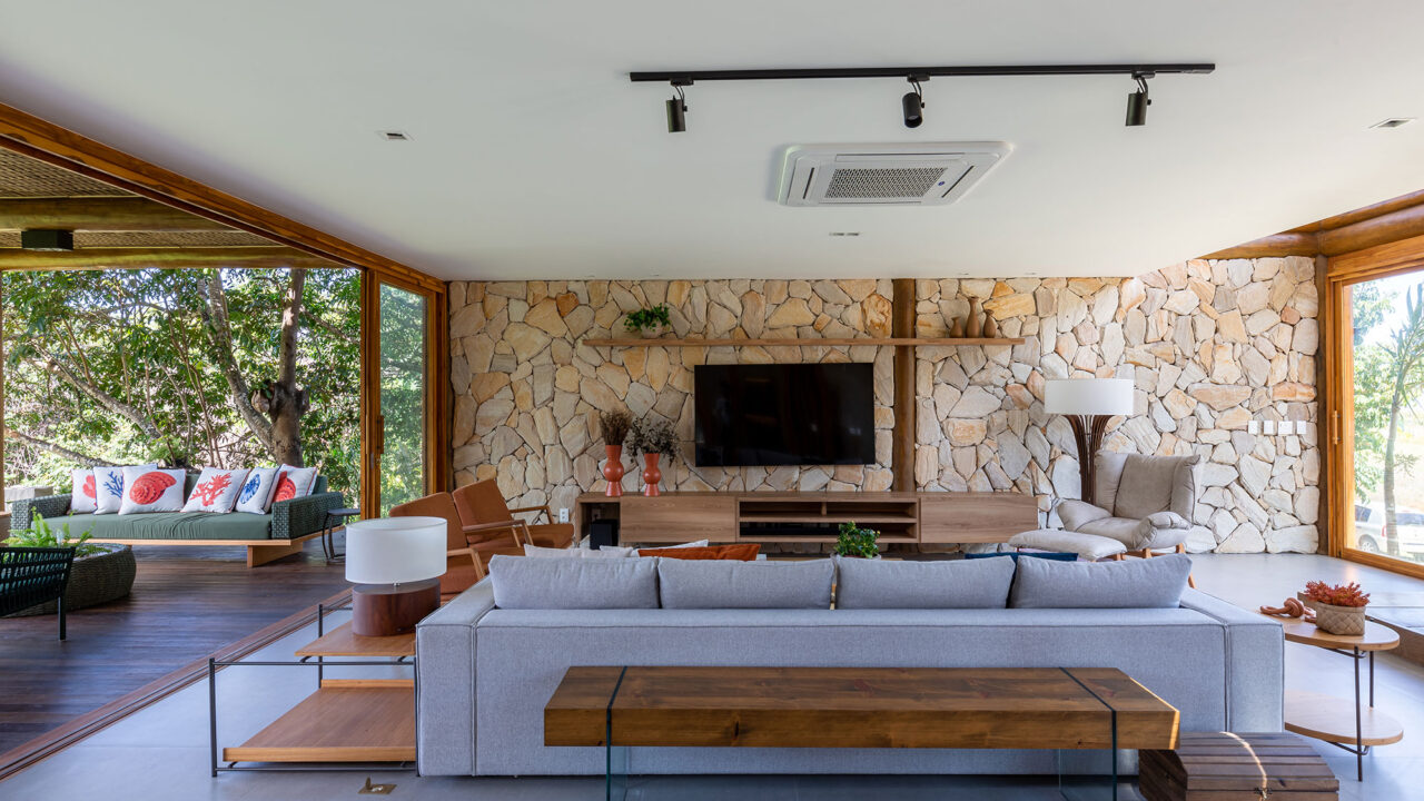 Casa na praia se integra à natureza com venezianas móveis na fachada. Projeto de Sidney Quintela. Na foto, sala de tv com parede de pedra e varanda.