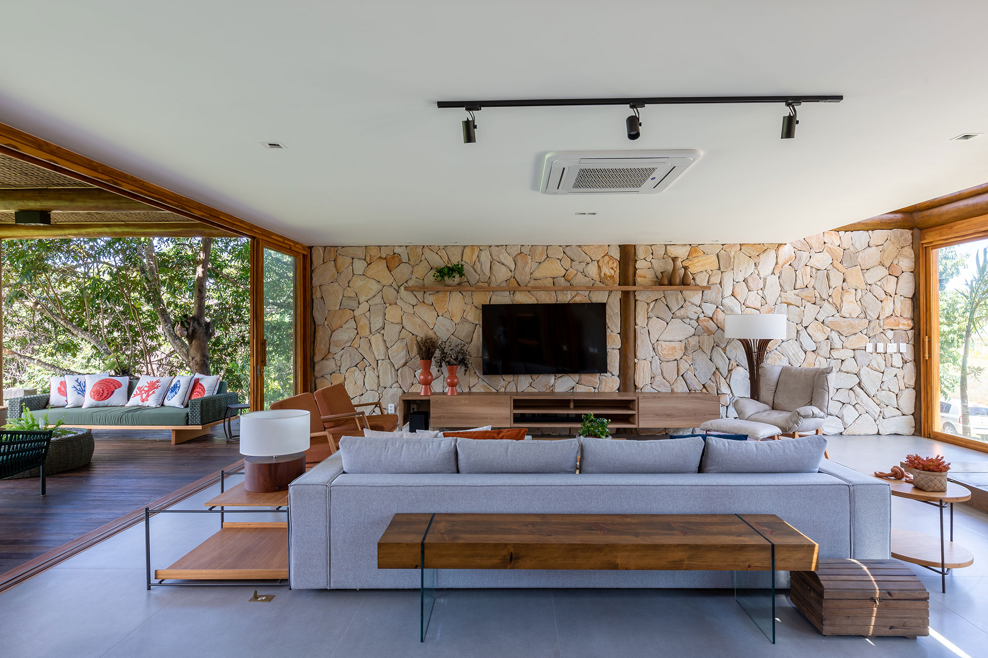 Casa na praia se integra à natureza com venezianas móveis na fachada. Projeto de Sidney Quintela. Na foto, sala de tv com parede de pedra e varanda.