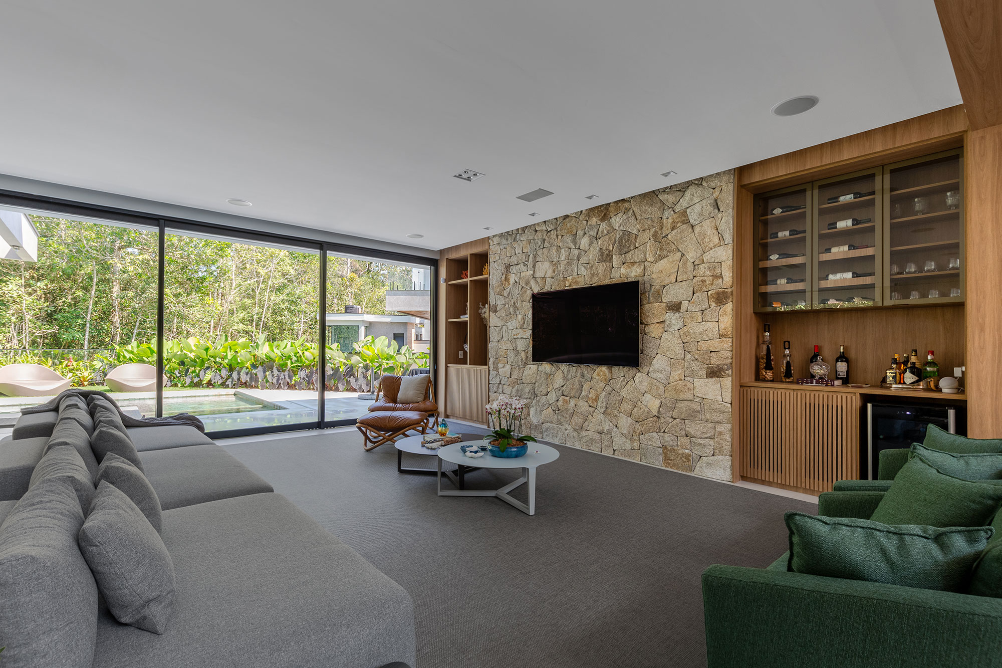 Com estilo rústico, casa de praia é um refúgio em meio ao verde. Projeto de Sabrina Salles. Na foto, sala com bar, parede de pedra e TV.